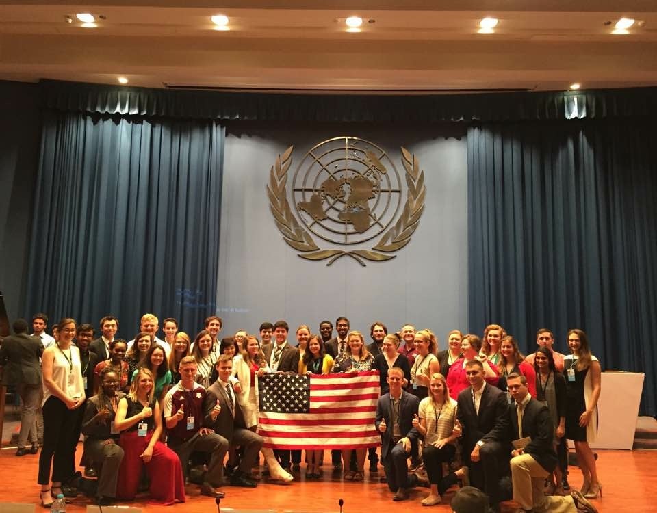 A large group photo on a stage holding the U.S. flag. Behind, a large logo of the UN